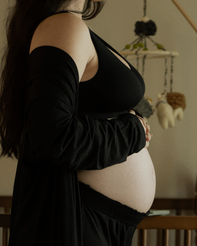 A pregnant woman wearing a black maternity top and a long black robe, standing in a softly lit indoor setting with a crib and hanging nursery decor in the background.
