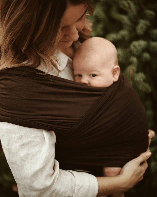 A woman wearing a white shirt holds a baby in a dark brown wrap-style baby carrier outdoors, with soft-focus green foliage in the background.