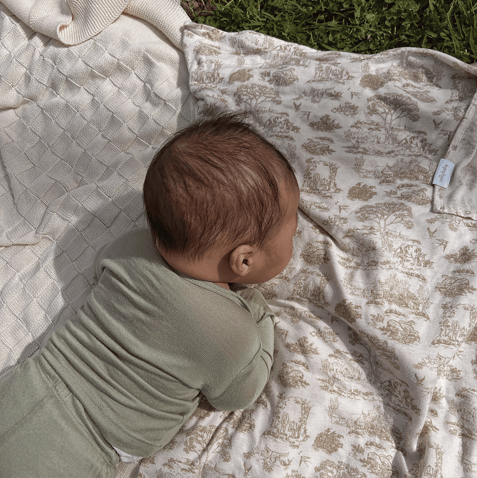 Baby resting on a soft Solly Baby blanket featuring the delicate Toile design in warm cream and taupe tones. The baby is dressed in a cozy sage green outfit, lying on a textured knit blanket and toile fabric. A peaceful outdoor moment wrapped in softness and comfort.