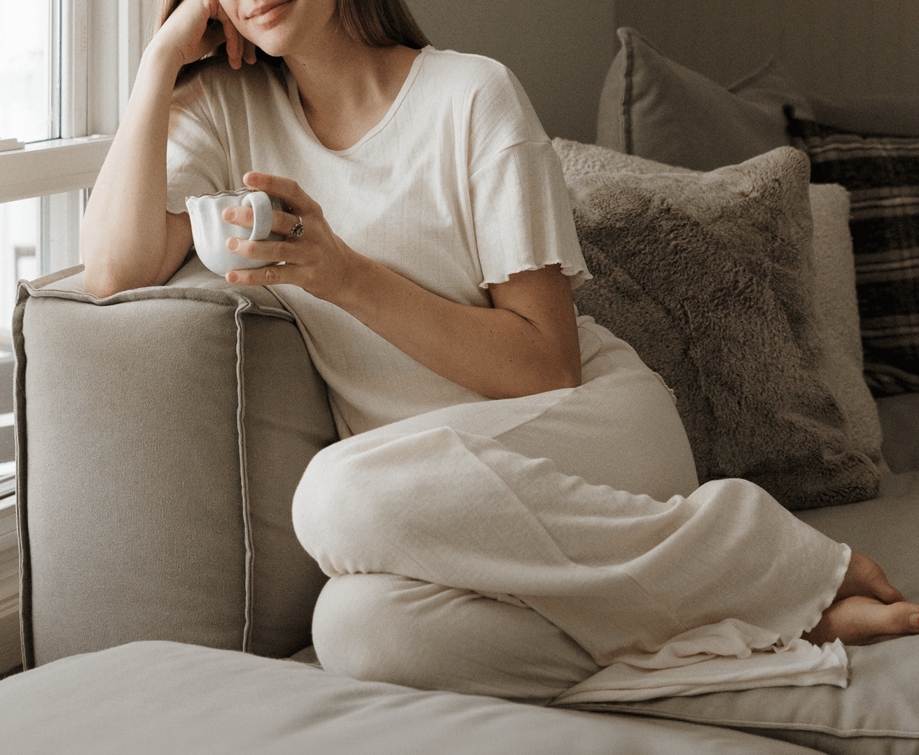 Mom sitting on a couch holding a mug, waring a cozy cream pointelle matching women's sleep set with lettuce edge hems