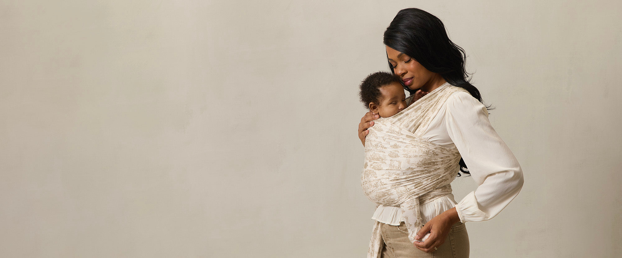 A mother holds her sleeping baby close in a Solly Baby Wrap featuring a delicate cream and soft beige toile print. The wrap provides a snug, secure hold, allowing the baby to rest peacefully against her chest. She pairs the wrap with a flowing cream blouse and soft-toned pants, creating an elegant and serene look.