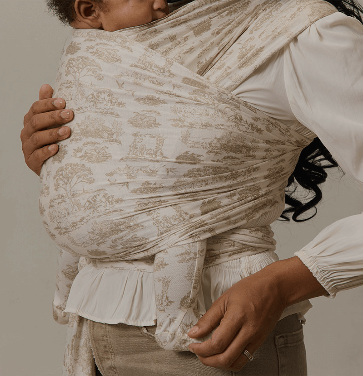 A mother holds her sleeping baby close in a Solly Baby Wrap featuring a delicate cream and soft beige toile print. The wrap provides a snug, secure hold, allowing the baby to rest peacefully against her chest. She pairs the wrap with a flowing cream blouse and soft-toned pants, creating an elegant and serene look.