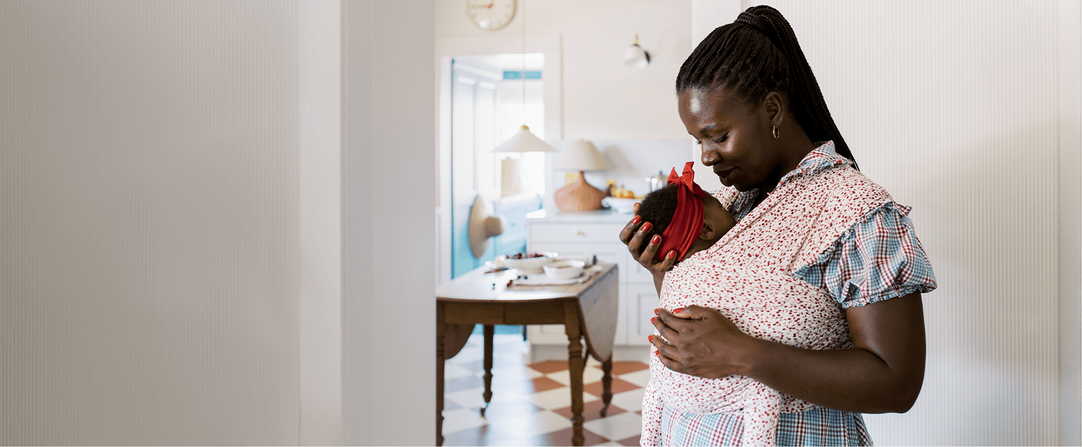 Mother stands in a cozy kitchen cradling her baby in the Solly Baby Cherry Wrap, radiating tenderness and calm. A beautiful moment of everyday connection during summer at home.