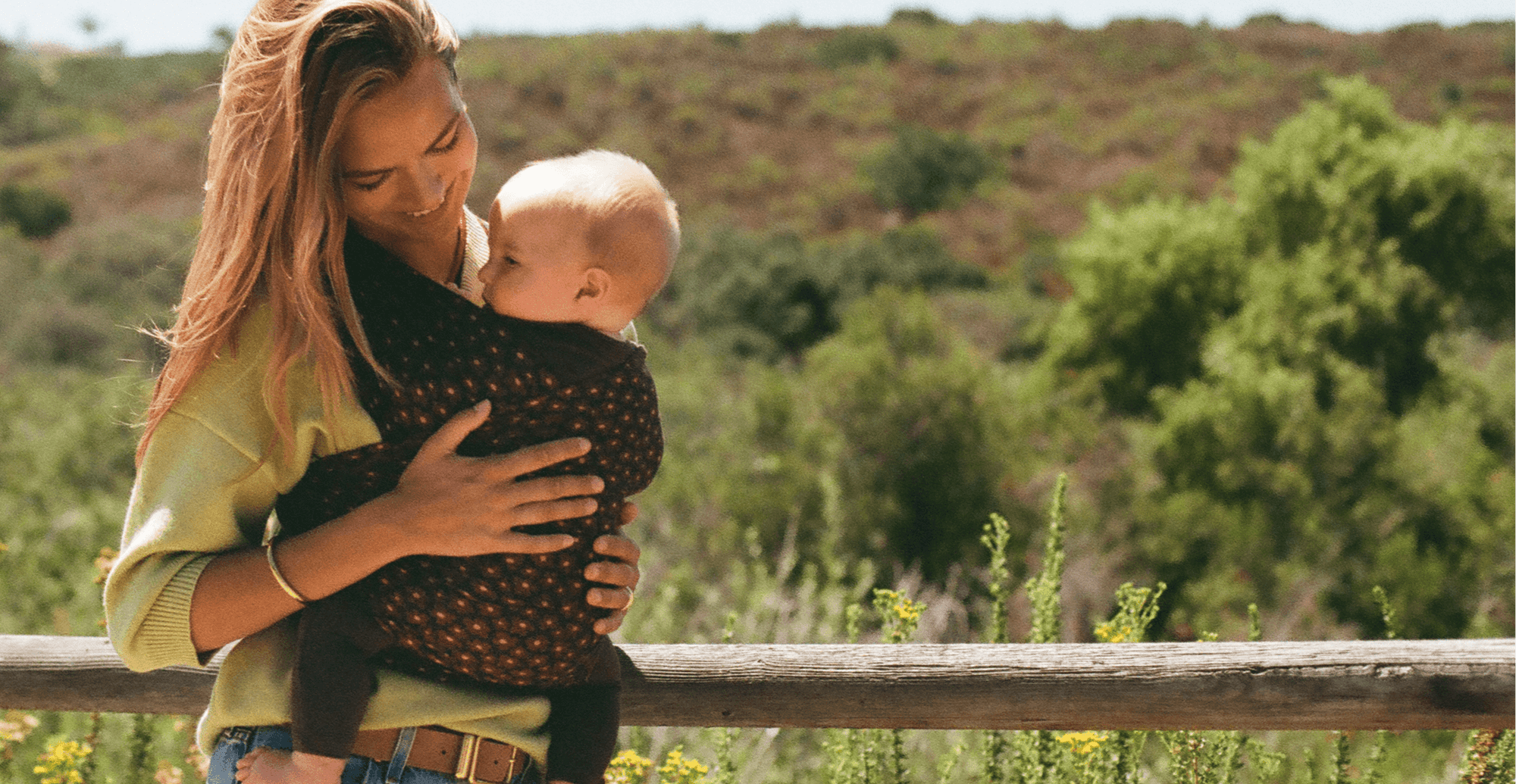 A woman outdoors wearing a dark patterned baby wrap carrier with a baby inside, set against a blurred natural background of green trees and hills.