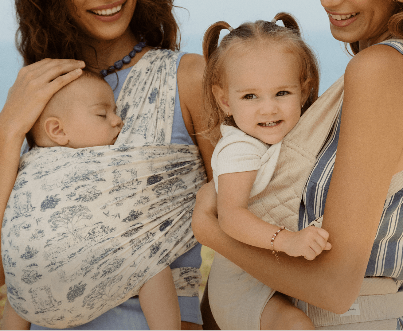 A close-up lifestyle shot of a mother and daughter wearing baby carrier wraps. The mother on the left wears a light blue dress and holds a baby in a white wrap with a blue floral print.
