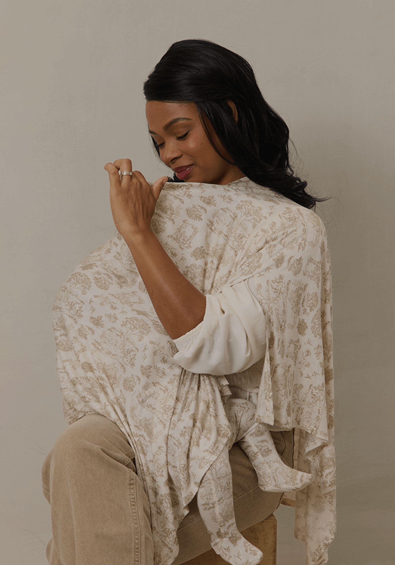 A mother sits comfortably while feeding her baby under a Solly Baby Nursing Cover in a delicate cream and beige toile print. The lightweight cover drapes elegantly over her shoulders, offering privacy and ease while the baby’s tiny feet peek out beneath the fabric.