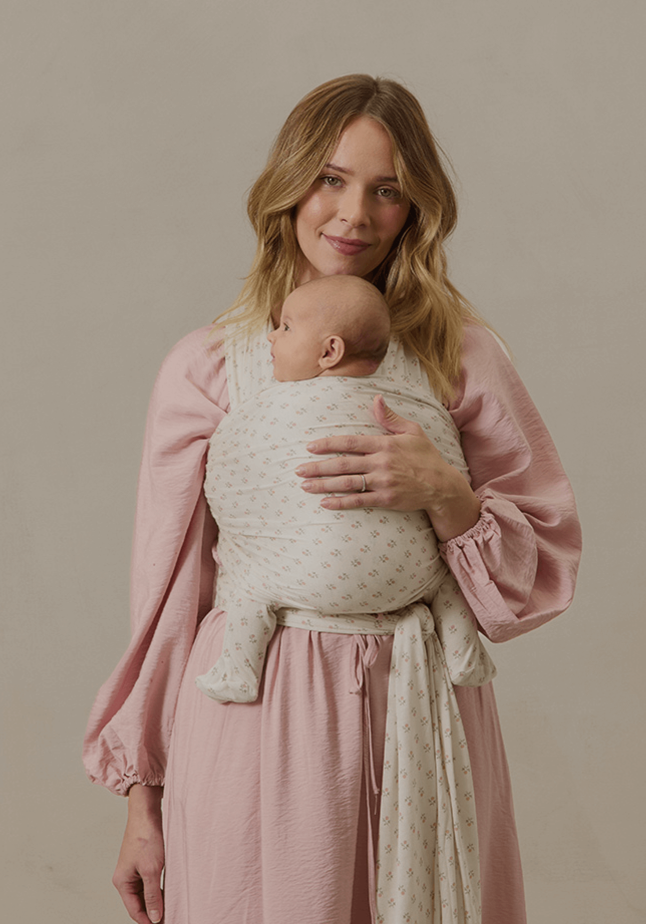 A mother cradles her baby in a Solly Baby Wrap featuring a soft cream base with a subtle floral pattern. She wears a flowing pink dress, and the wrap is tied securely, providing a snug, cozy hold as the baby rests contentedly against her chest.