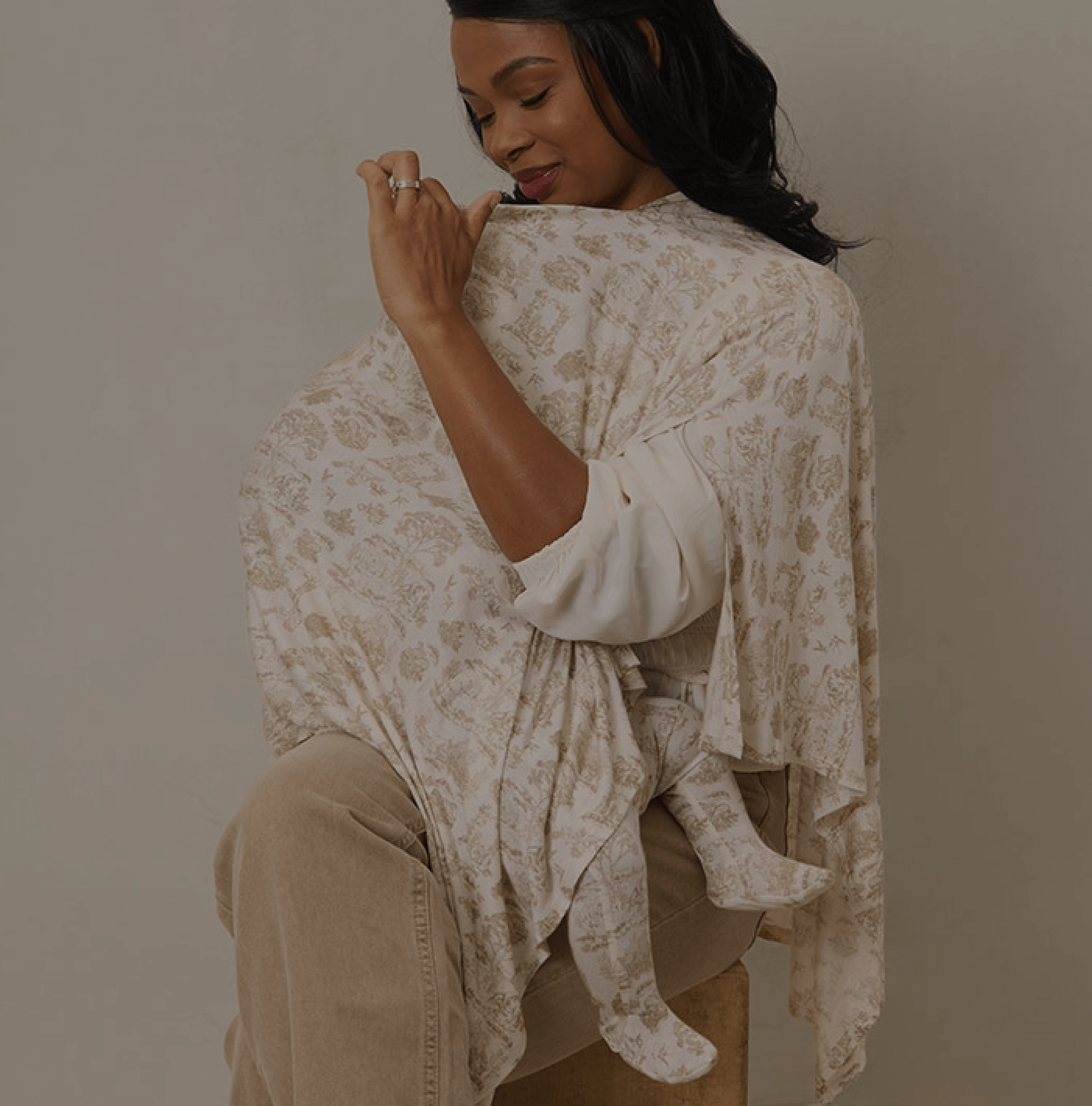 Mother nursing her baby under a Solly Baby nursing cover featuring the delicate Winter Toile design in warm cream and taupe tones. The baby's tiny feet peek out from beneath the soft, breathable fabric, creating a cozy and comforting moment for both mother and child.