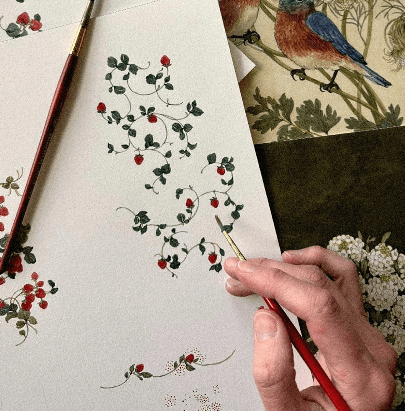 Hand painting intricate strawberry designs on a canvas with a brush, surrounded by nature-themed artwork.