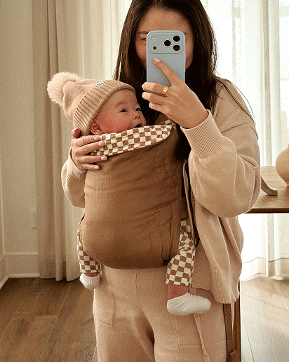 A woman takes a mirror selfie in a bright room while wearing a beige cardigan and matching pants, holding a baby in a brown front baby carrier. The baby wears a beige and white checkered hat and matching checkered foot coverings.