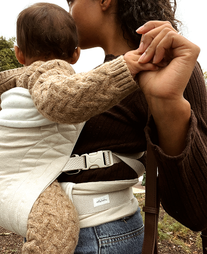 A close-up lifestyle shot of a woman wearing a brown ribbed knit baby carrier with a baby in a cream quilted insert. The woman is wearing a dark brown sweater and blue jeans, and the baby is wearing a beige cable-knit sweater.