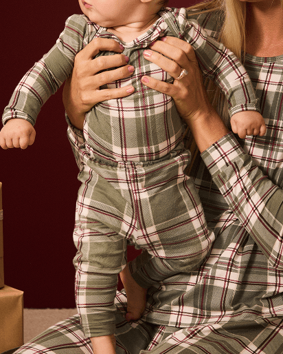 A baby wearing a matching plaid romper and pants set, held by an adult wearing a similar long-sleeve top. The fabric has an olive green base with a red and cream plaid pattern, set against a dark burgundy background.