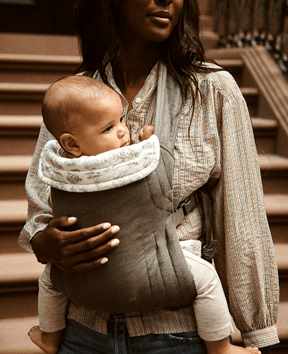 A woman wearing a striped button-up shirt carries a baby in a dark gray quilted baby carrier wrap with a white patterned bib insert, set against a blurred background of outdoor stone steps.