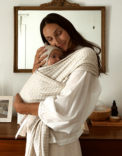 A woman wearing a white blouse holds a baby wrapped in a cream and beige checkered baby wrap carrier, standing in front of a mirror in a warm, neutral-toned room.