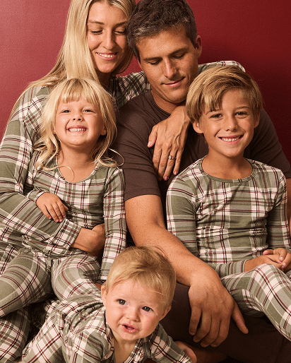 A family of five wearing matching green and brown plaid long-sleeve pajamas, posed together against a solid dark red background.