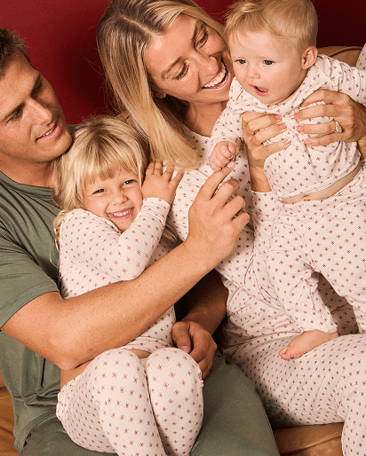 A family of four wearing matching red and white twinkle star patterned pajamas, hugging together against a solid red background.