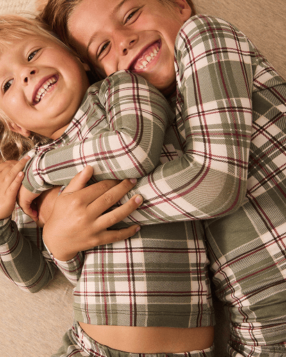 Two children hugging while wearing matching long-sleeve plaid pajamas in muted green, cream, and burgundy tones, lying on a beige carpet.