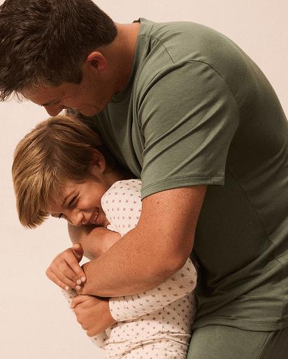 A man in an olive green t-shirt and matching pants hugging a young child wearing a white patterned long-sleeve top against a plain light beige background.
