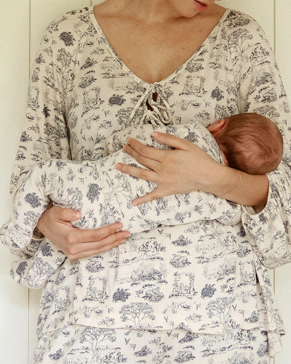 A woman wearing a matching cream and gray toile-print babywearing outfit, cradling a baby wrapped in the same patterned swaddle.