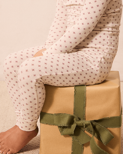 A person wearing cream pajama pants with a small red star pattern sits barefoot on a wrapped gift box. The photo is cropped from the waist down against a plain beige background.