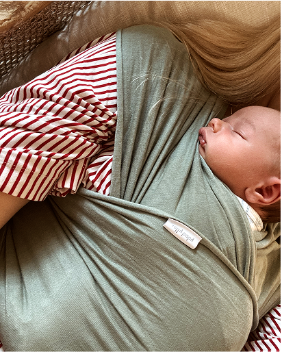 A sleeping newborn baby is wrapped in a sage green baby wrap carrier, resting on a person's chest. The person is wearing a red and white striped shirt.