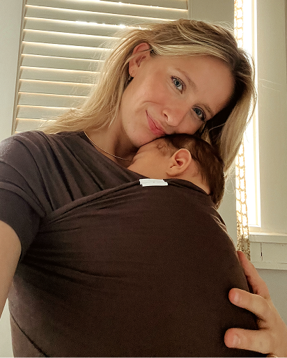 A woman wearing a dark brown baby wrap carrier with her baby, in a close-up selfie-style photo in front of window blinds.