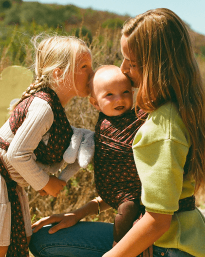 Three women outdoors wearing matching dark red patterned baby wraps over white and lime green tops, with a baby in the center wrap and another woman kissing the baby on the cheek.