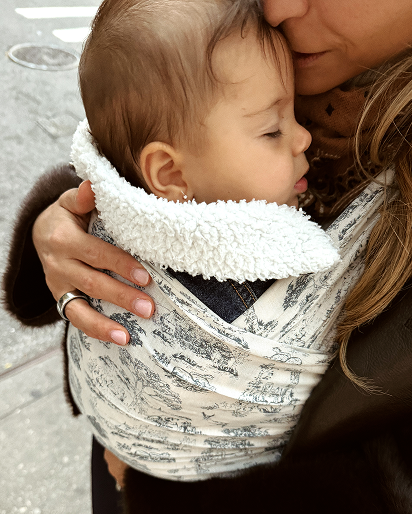A close-up lifestyle shot of a baby in a wrap carrier worn by a mother. The carrier is made of a white fabric with a gray toile de Jouy pattern and features a thick, plush white sherpa lining.