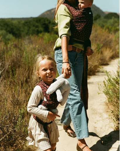 A woman and a young girl walking on a dirt trail outdoors, each wearing a patterned baby carrier wrap. The scene is set in a natural, grassy landscape with hills in the background.
