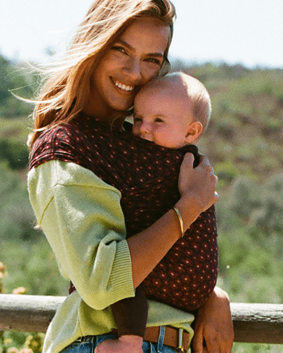 A woman outdoors wearing a dark pink baby carrier wrap with a floral pattern, carrying a baby. She is also wearing a light green knit top.