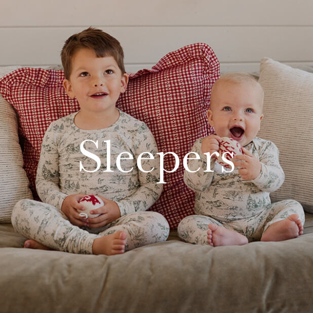 Two children in matching cream and green winter toile pajamas holding ornaments while sitting on a couch with red pillows. Shop Sleepers. 