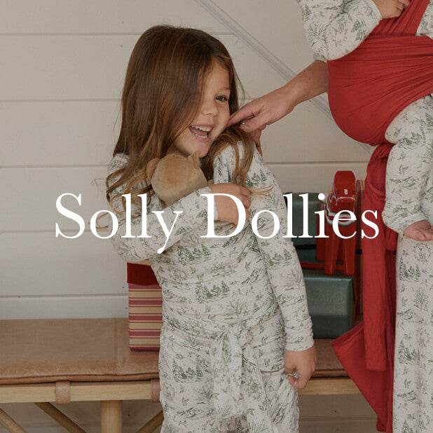 Child smiling and holding a doll in a solly dolly wrap while wearing cream and green winter toile pajamas, standing near wrapped holiday gifts. Shop Solly Dollies. 