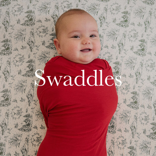 Smiling baby swaddled in a vibrant red wrap lying on a crib sheet with a green winter toile design. Shop Swaddles. 