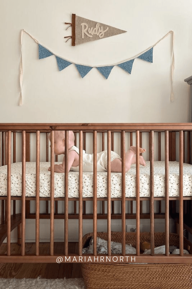 Baby in a crib with strawberry printed sheet and a decorative banner above, featuring the name 'Rudy'.