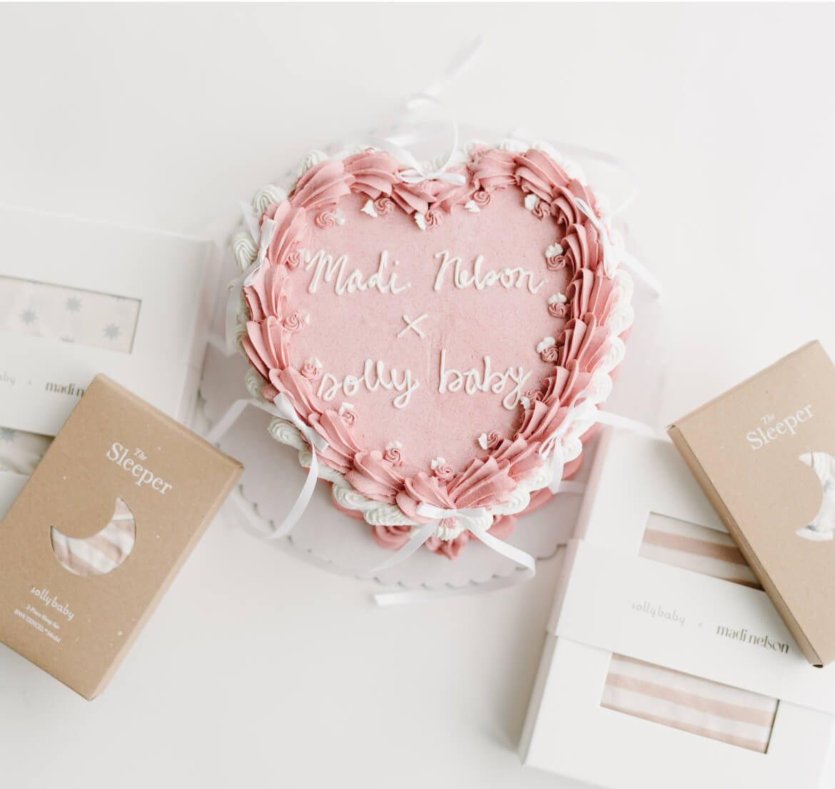 Flat lay showcasing the Solly Baby x Madi Nelson collection, featuring cozy baby essentials like a striped sleeper in soft neutral tones, packaged in kraft boxes with crescent moon cutouts. A delicate pink heart-shaped cake with piped details reads 'Madi Nelson x Solly Baby,' surrounded by confetti accents. Celebrate the tender moments of motherhood with timeless, thoughtfully designed pieces.