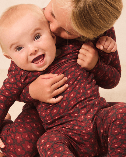 A baby and an adult wearing matching dark red patterned one-piece pajamas, with the adult kissing the baby's cheek in a warm indoor setting.