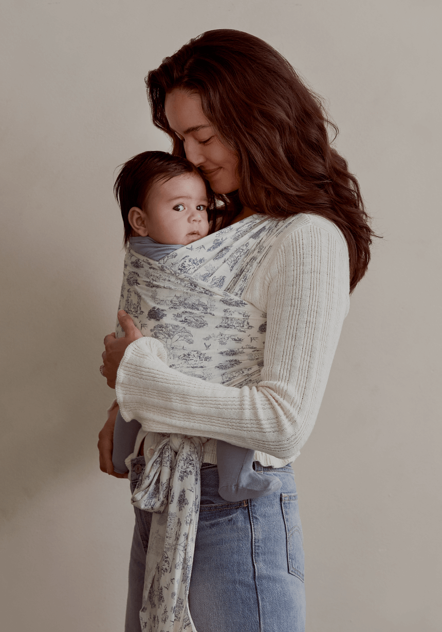 A mother cradles her baby in a Solly Baby Wrap featuring a cream and soft blue toile design. The wrap provides a snug, secure hold as the baby rests contentedly, dressed in coordinating slate blue. She pairs the wrap with a soft, cream knit sweater and relaxed jeans.