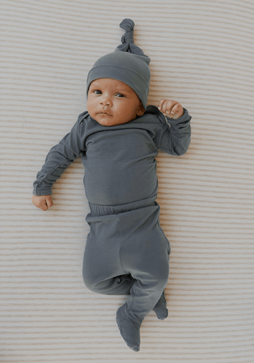 A newborn baby lies peacefully on a soft, neutral-striped surface, dressed in a matching slate blue set featuring a long-sleeve top, footed pants, and a knotted hat. The baby's gaze is calm and curious, showcasing comfort and style in the essentials for little ones.