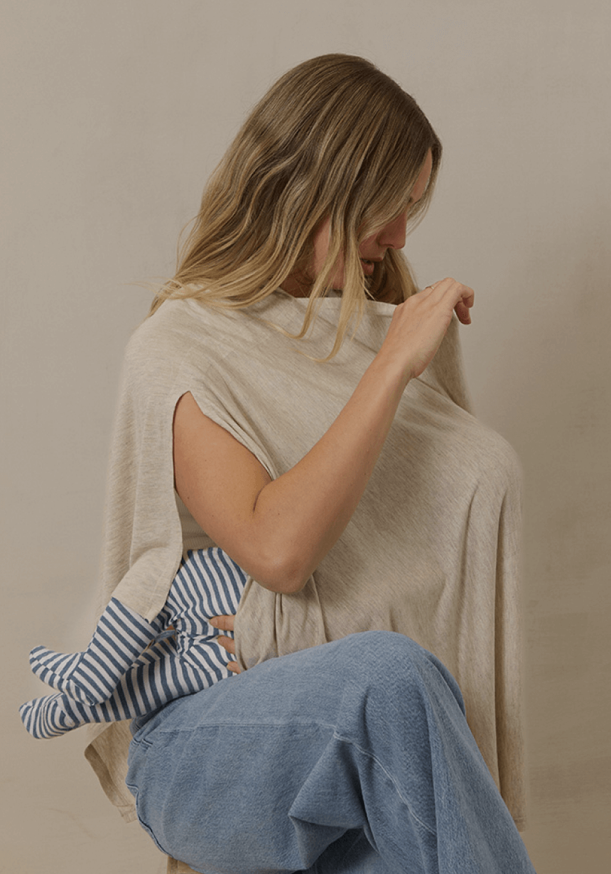 A mother sits comfortably while feeding her baby under a Solly Baby Nursing Cover in a soft, neutral tone. The lightweight fabric drapes gently over her shoulders, providing privacy and ease. The baby’s small feet, wrapped in a blue and white striped sleeper, peek out from beneath the cover.