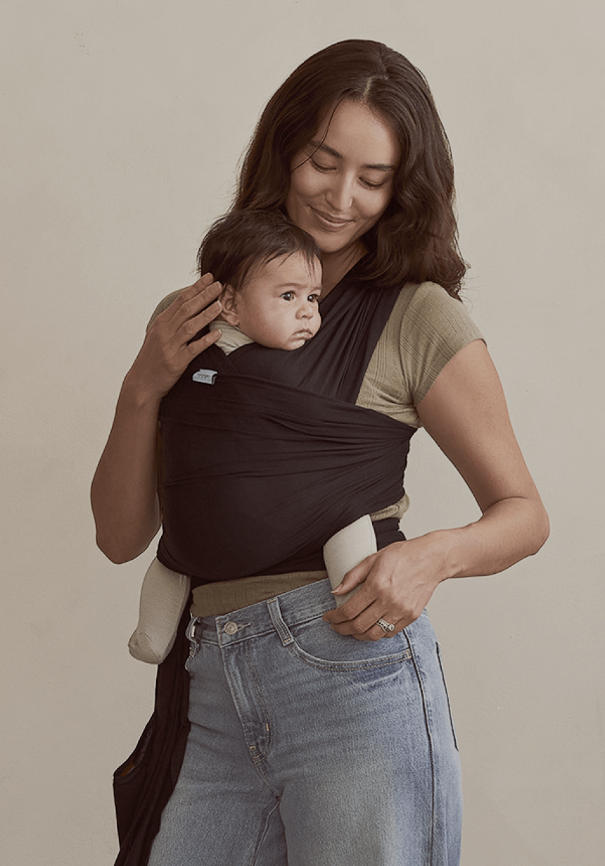 A mother holds her baby close in a black Solly Baby Wrap, creating a secure and cozy bond. The wrap drapes snugly around her as the baby looks outward, resting comfortably against her chest. Styled effortlessly with a neutral tee and relaxed denim.