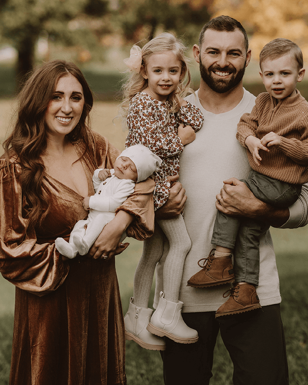 A family of four posing outdoors in warm, earthy tones. The mother wears a bronze-brown velvet dress and holds a baby in a white outfit.