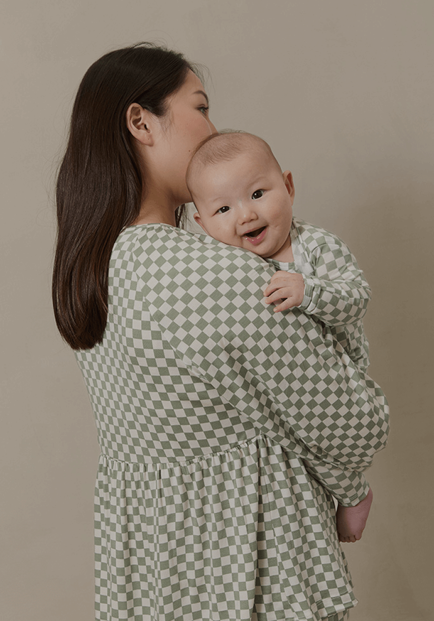 Mother holding her smiling baby, both dressed in Solly Baby matching sleepwear featuring a soft sage green checkerboard pattern. Cozy, breathable loungewear designed for sweet snuggles and restful nights.