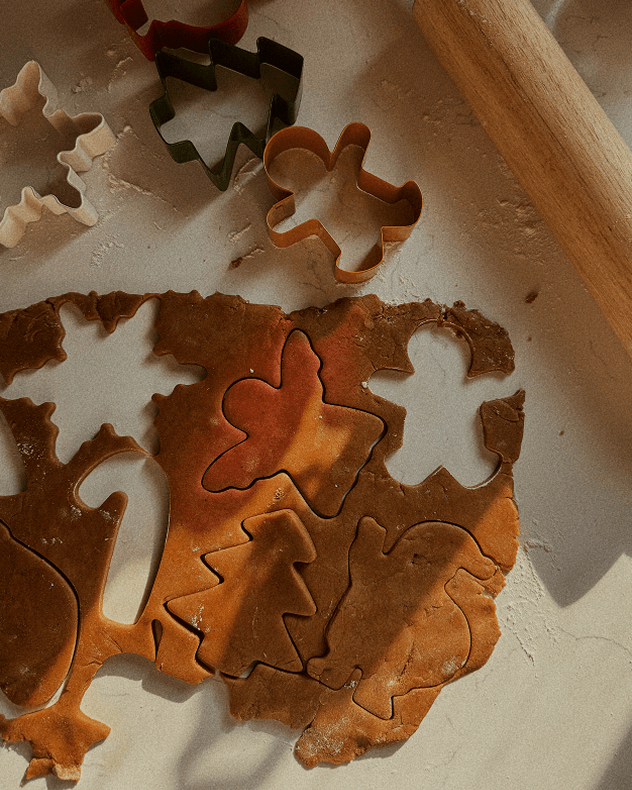 A close-up of holiday cookie cutters in various shapes, including gingerbread men, trees, and snowflakes, pressed into rolled-out gingerbread dough on a light surface with a wooden rolling pin visible.