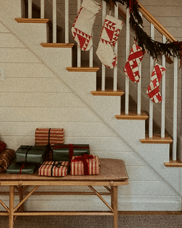 A styled holiday scene showing a wooden bench with wrapped Christmas presents underneath several cream and red patterned stockings hanging from a staircase with white railings and wood steps. A dark garland decorates the banister.