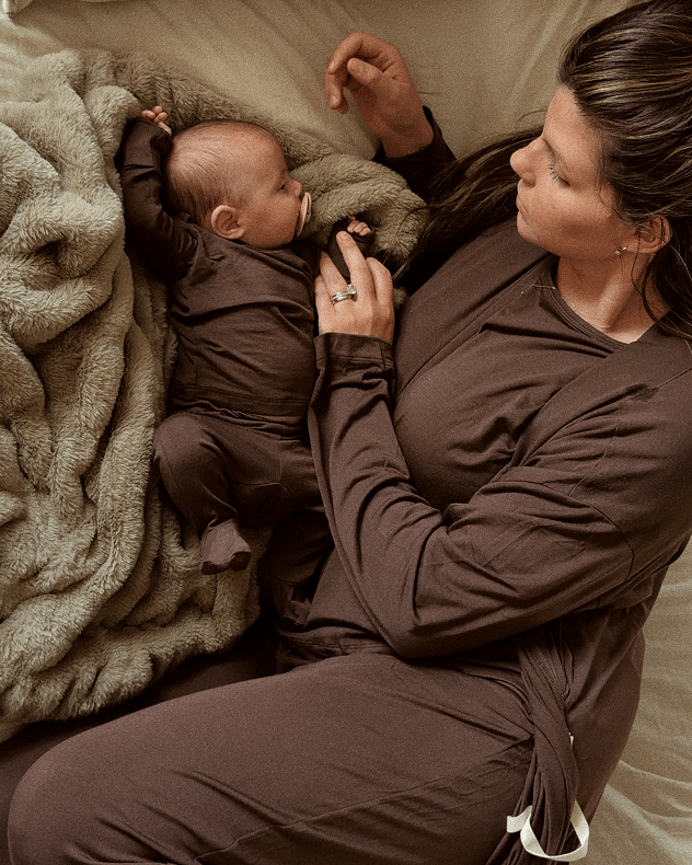 A woman and a baby wearing matching dark brown loungewear pajamas, lying on a beige bed next to a textured beige blanket.
