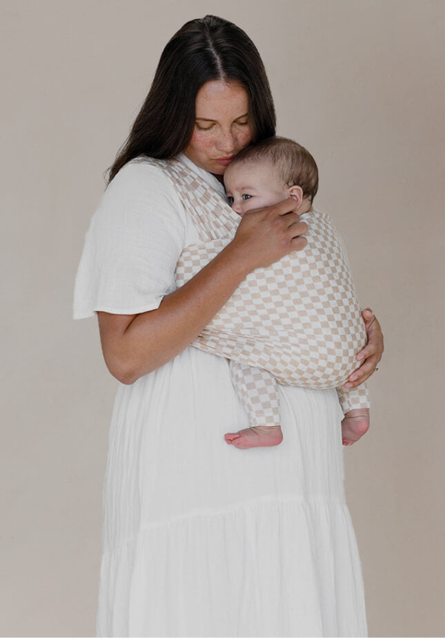 A mother standing while holding her baby in a Cream Chequer Solly Baby Wrap. 