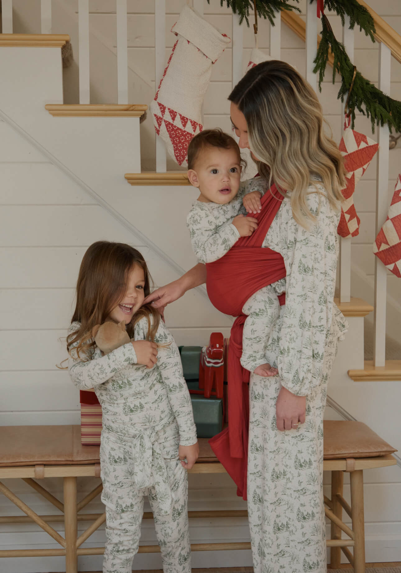 Family in matching cream and green winter toile pajamas, gathered by a holiday-decorated staircase.