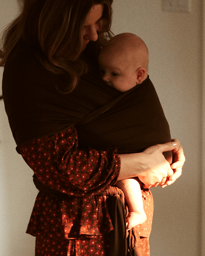 A woman wearing a dark brown wrap-style baby carrier holds a baby in a warm indoor setting. The carrier drapes over her shoulders and wraps around her torso and the baby.