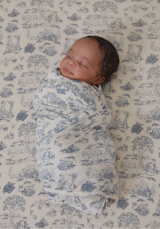 A newborn baby swaddled in Solly Baby Bleu Toile Wrap, lying on matching Bleu Toile Crib Sheet. 