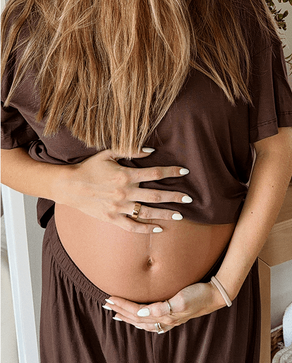 A close-up lifestyle shot of a pregnant woman wearing a dark brown short-sleeve maternity top and matching brown pants, cradling her baby bump with white manicured nails.